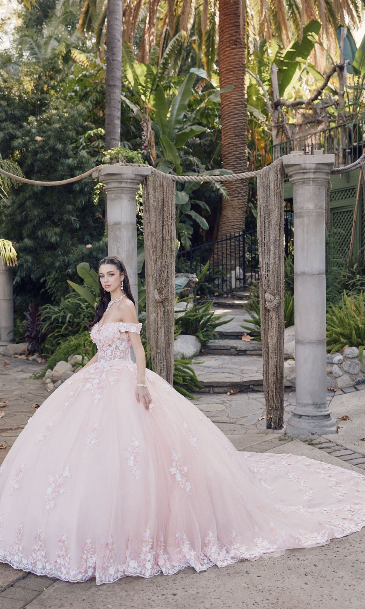 A young woman wears the Quinceanera Dress By Juliet JT1460J, an off-the-shoulder ball gown with floral details, as she stands outdoors between stone columns amid lush greenery and palm trees.